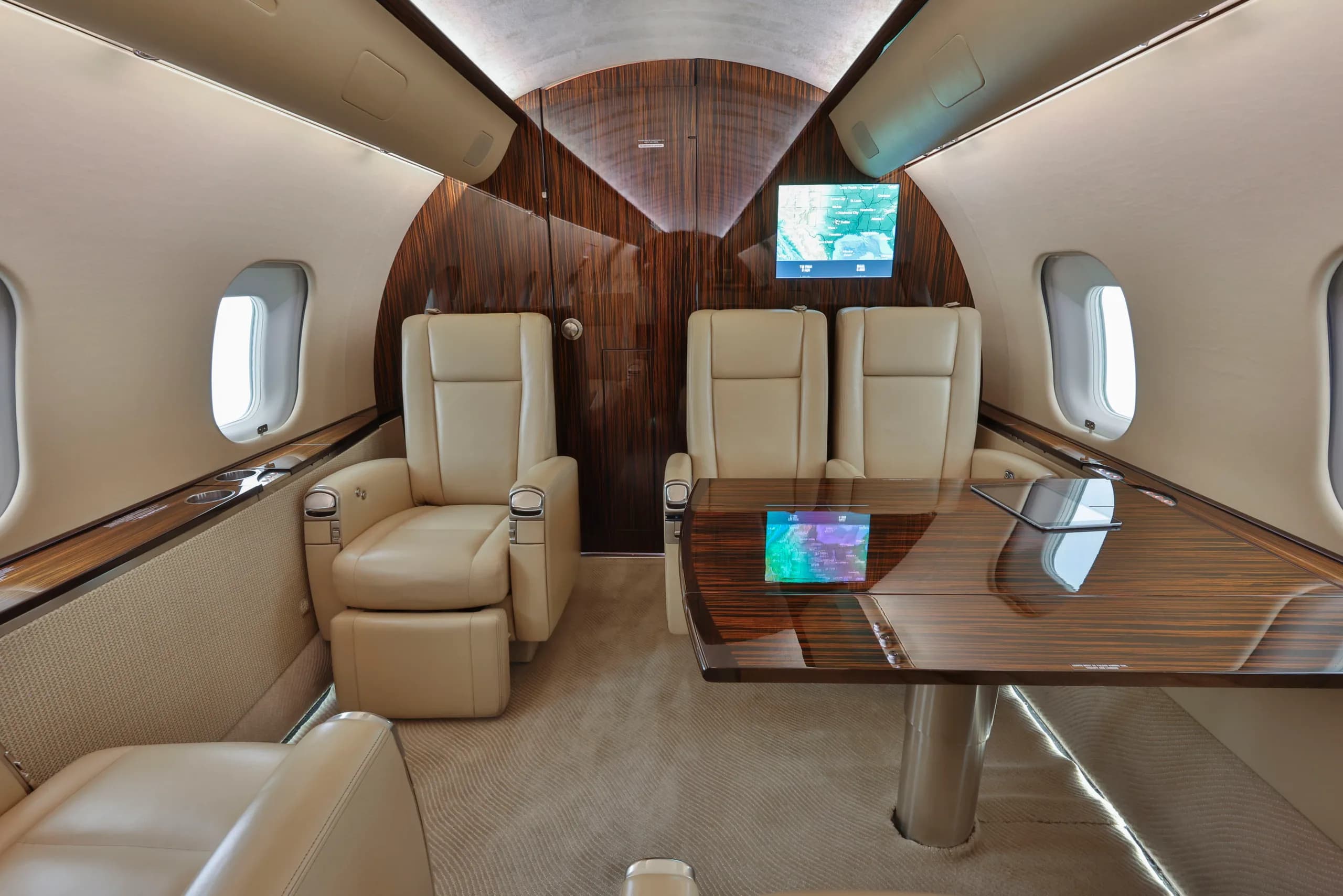 A section of the business jet's cabin with two beige leather seats, a wooden table, and two wall-mounted screens.