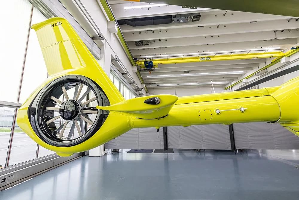 A close-up view of the bright yellow tail boom and Fenestron tail rotor of an Airbus H135 helicopter in a hangar.