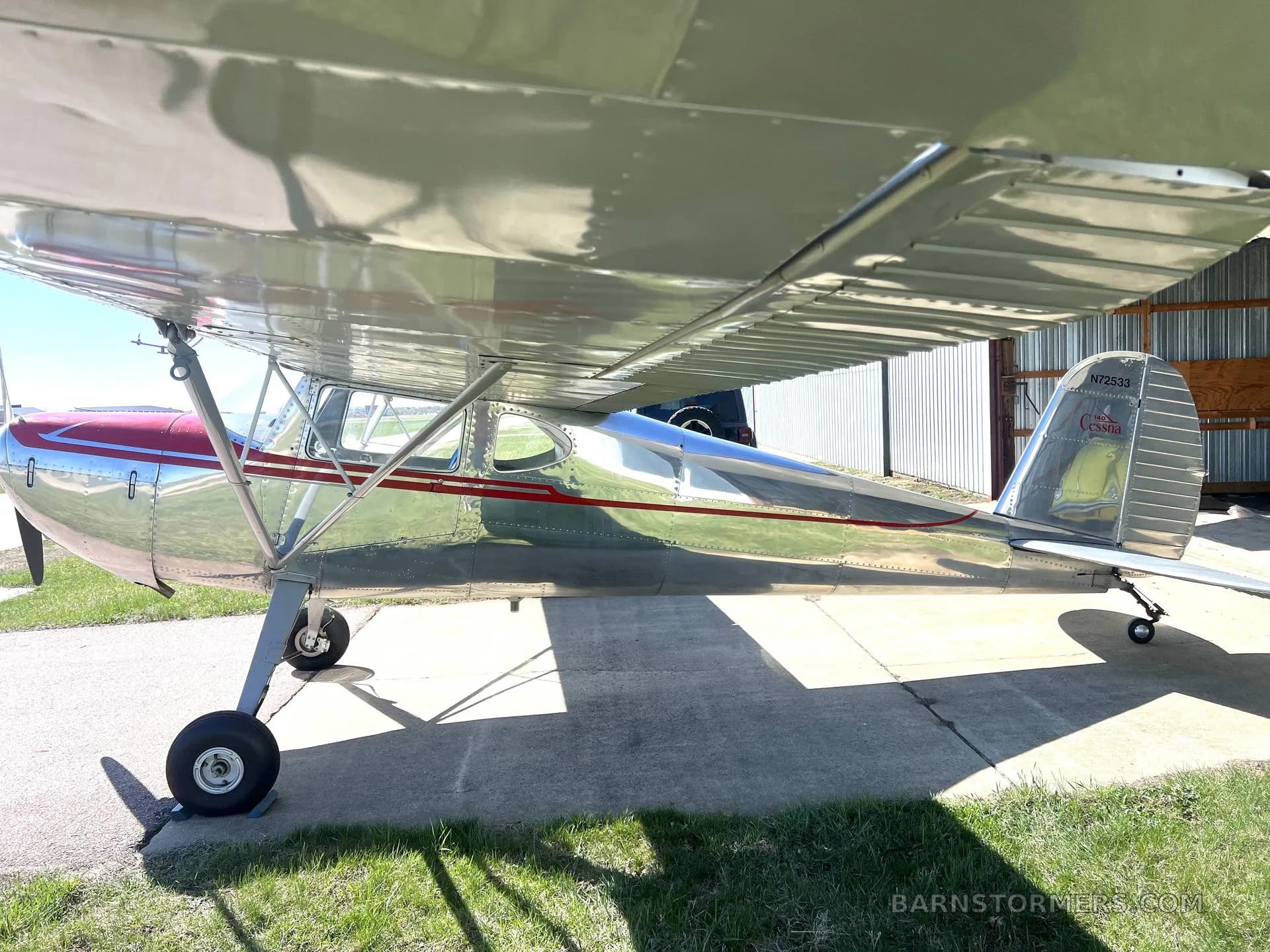 Side view of a silver Cessna 140 with red accents, showing the registration N72533 on the tail, parked next to a hangar.
