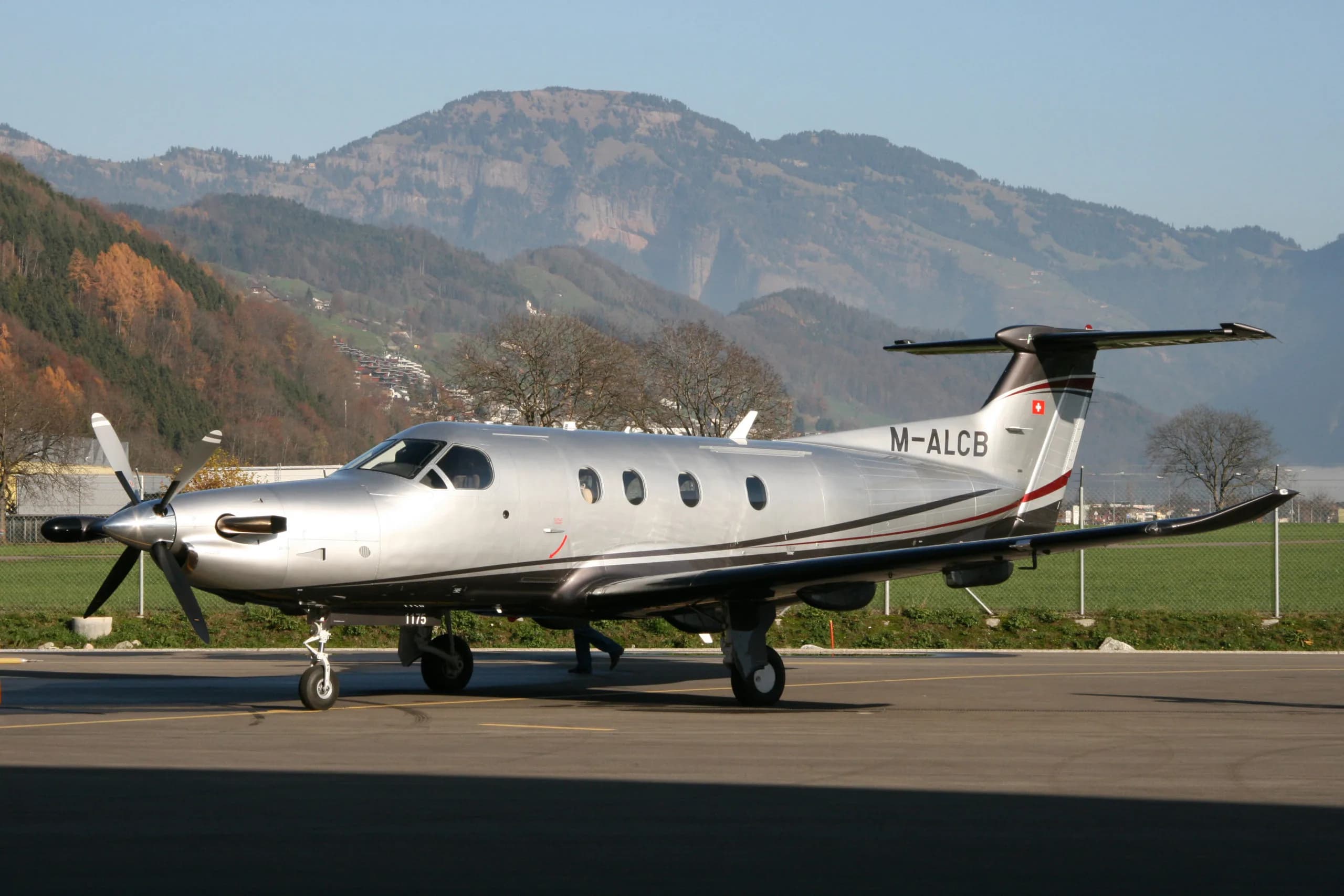 A silver Pilatus PC-12 aircraft with dark grey and red stripes is parked on an airfield with mountains in the background.