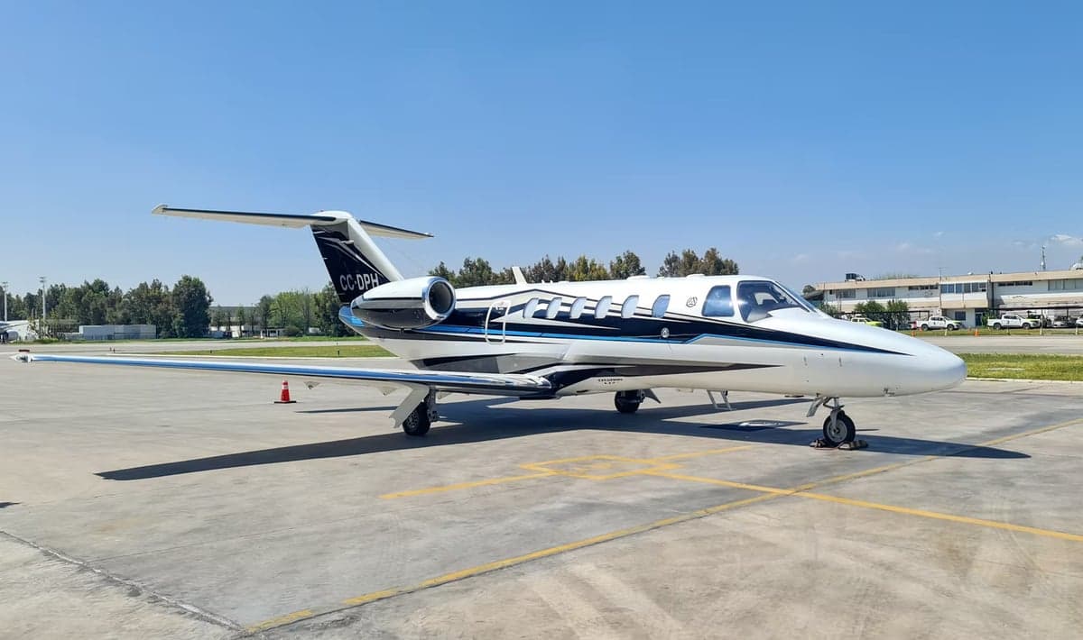 A white Cessna Citation Mustang jet with blue and black stripes is parked on the tarmac.