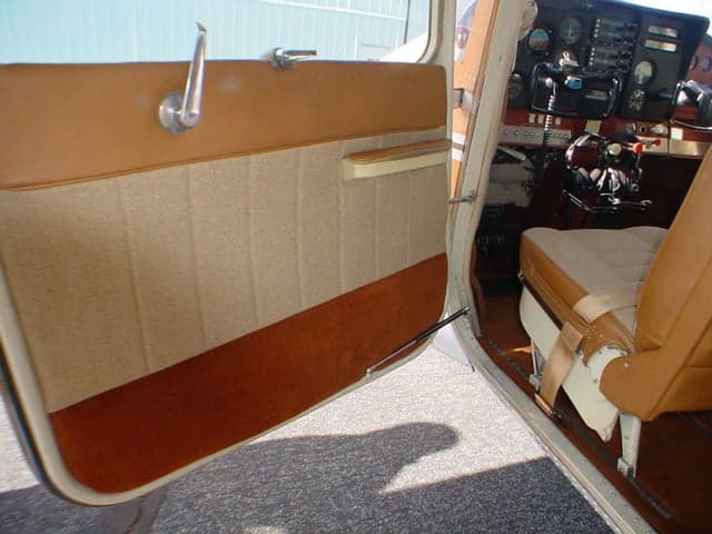 An open aircraft cabin door with tan and brown fabric trim, offering a view into the interior and partial cockpit.