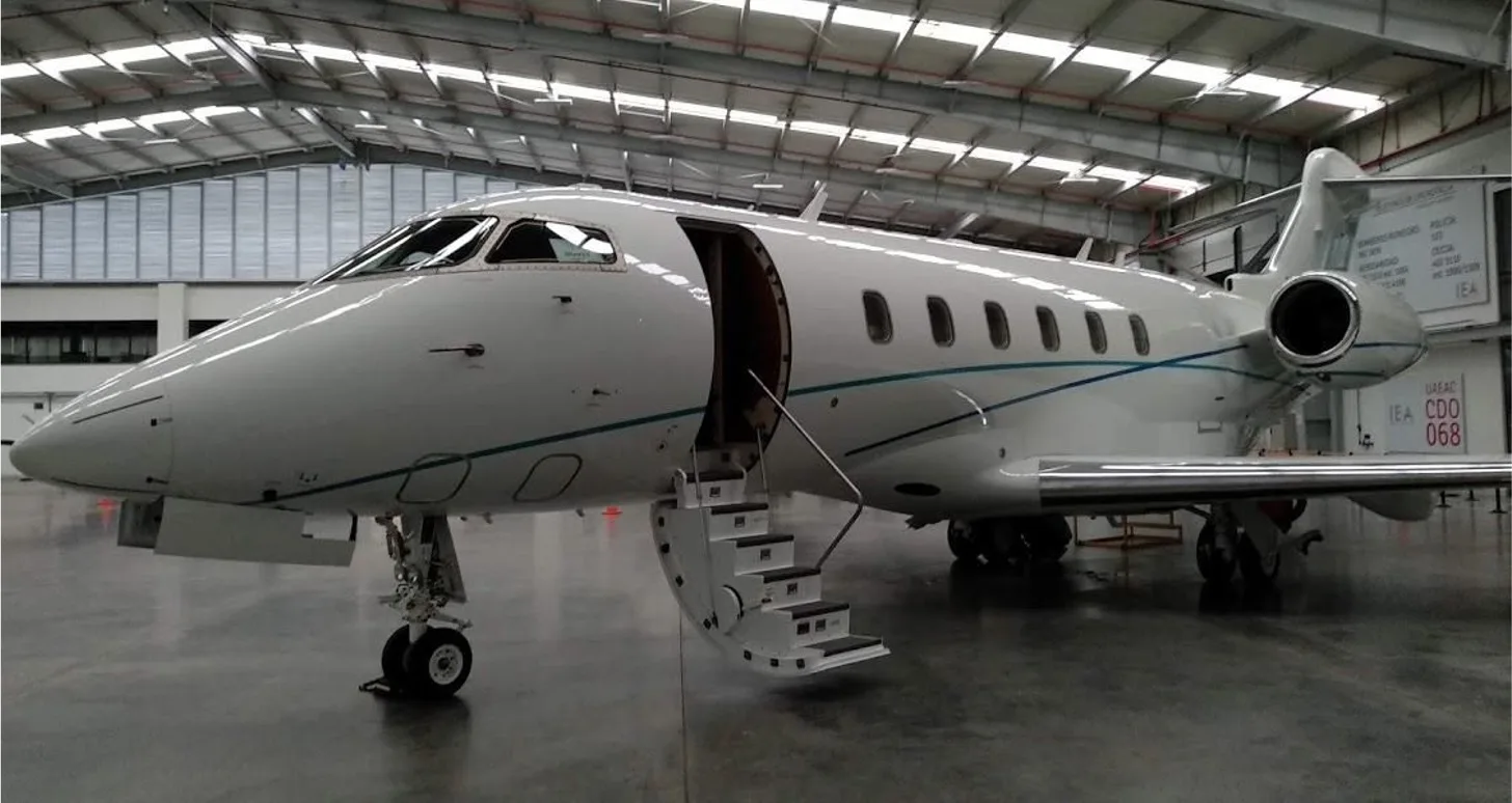 A white business jet with blue and teal stripes, registration "IEA", is parked in a hangar with its main cabin door open and airstairs extended.