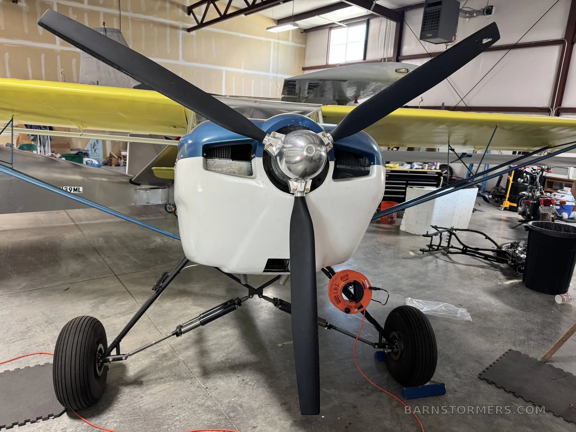 Front view of a yellow and blue Kitfox aircraft with a three-blade propeller, parked in a hangar.