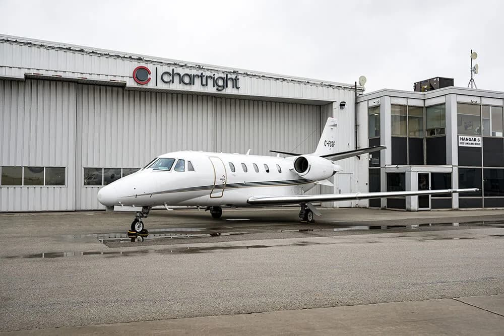 A white Cessna Citation business jet with a black stripe is parked on an asphalt apron in front of a Chartright hangar.