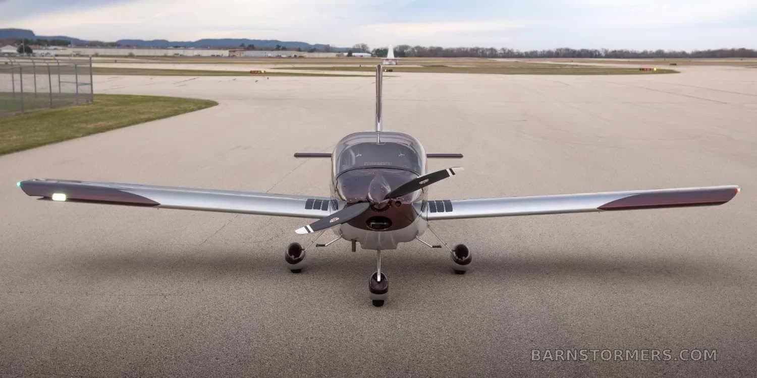 A head-on view of a maroon and silver single-engine propeller aircraft parked on an airfield with its landing lights on.