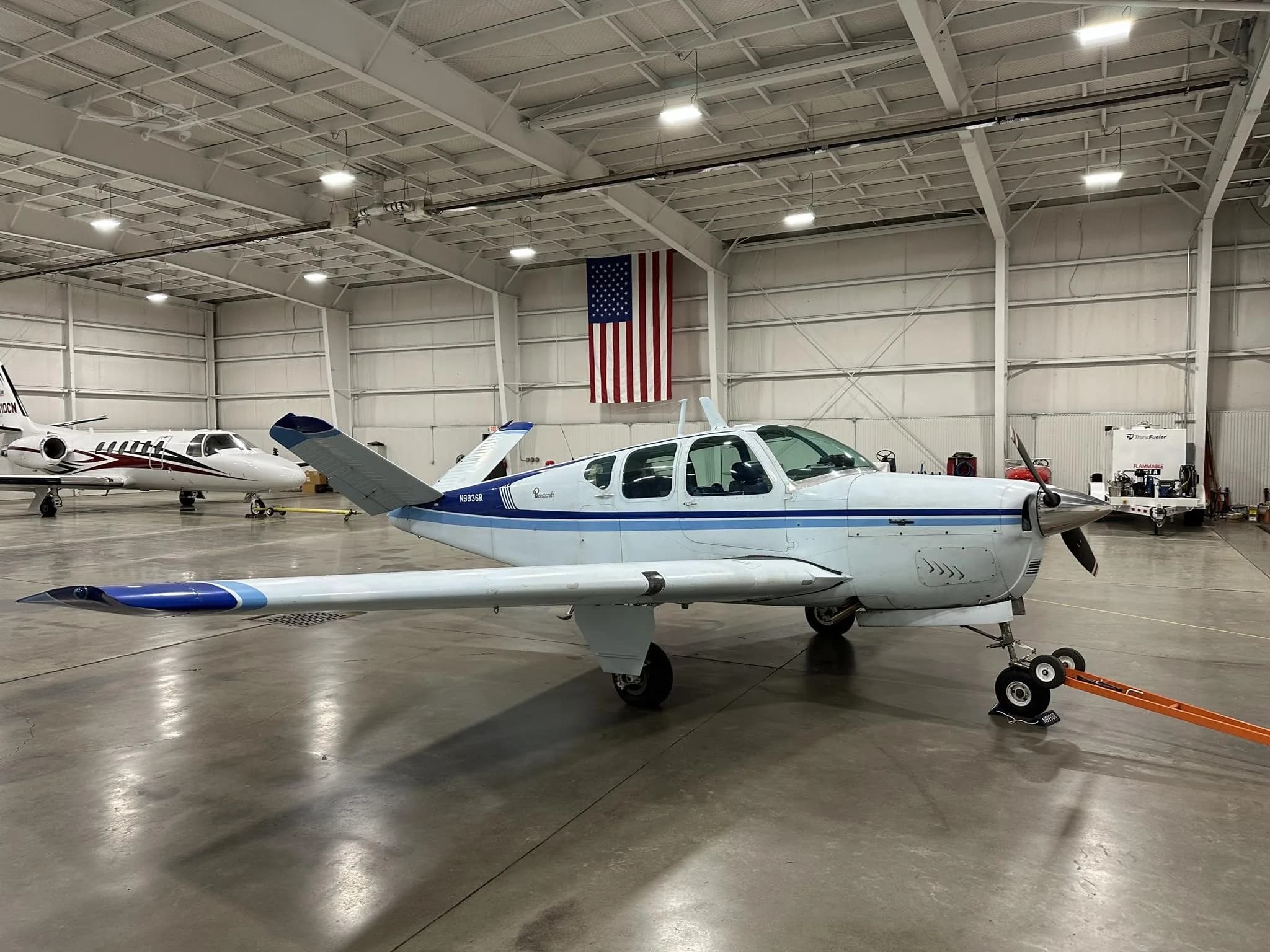 A white and blue Beechcraft Bonanza N9936R parked in a hangar with an American flag on the wall.