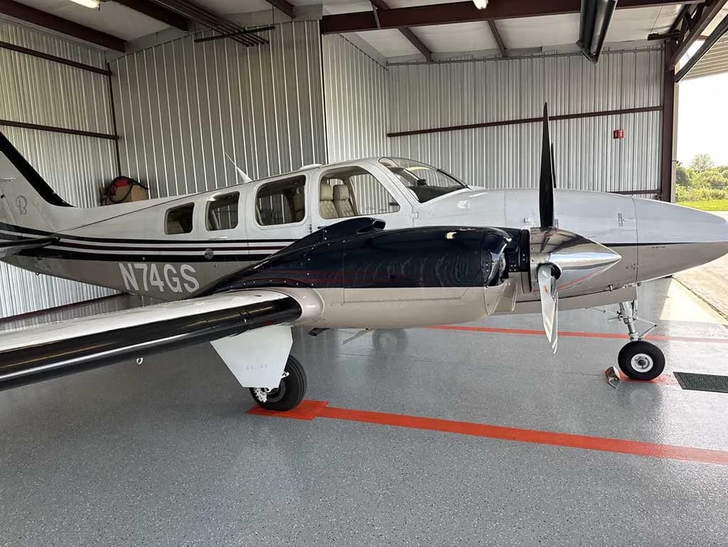 The white and tan twin-engine aircraft N74GS is parked inside a hangar with the door open to an airfield.