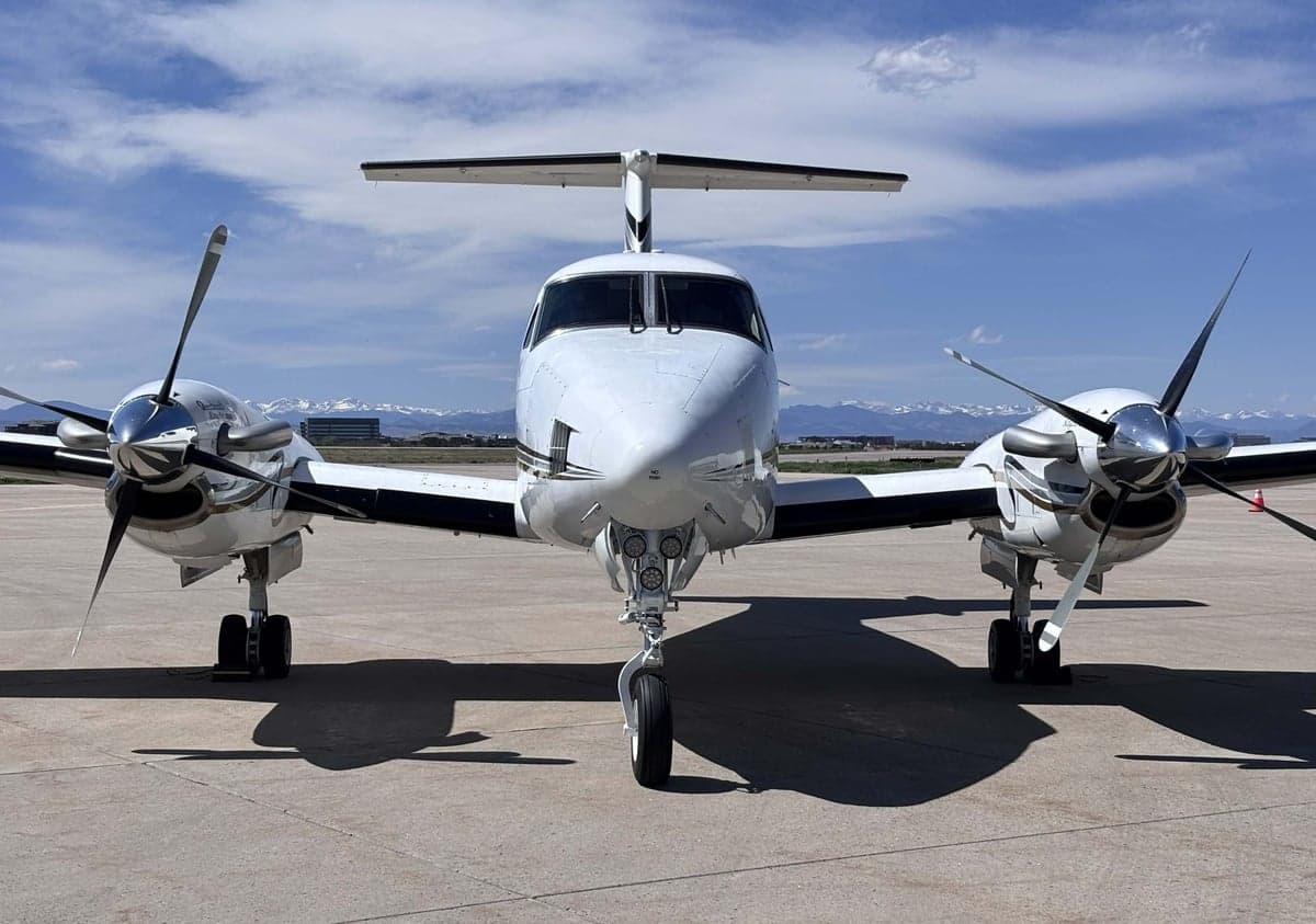 Front view of a white Beechcraft King Air N31WC with its propellers and landing gear visible, parked on an airfield with mountains in the background.