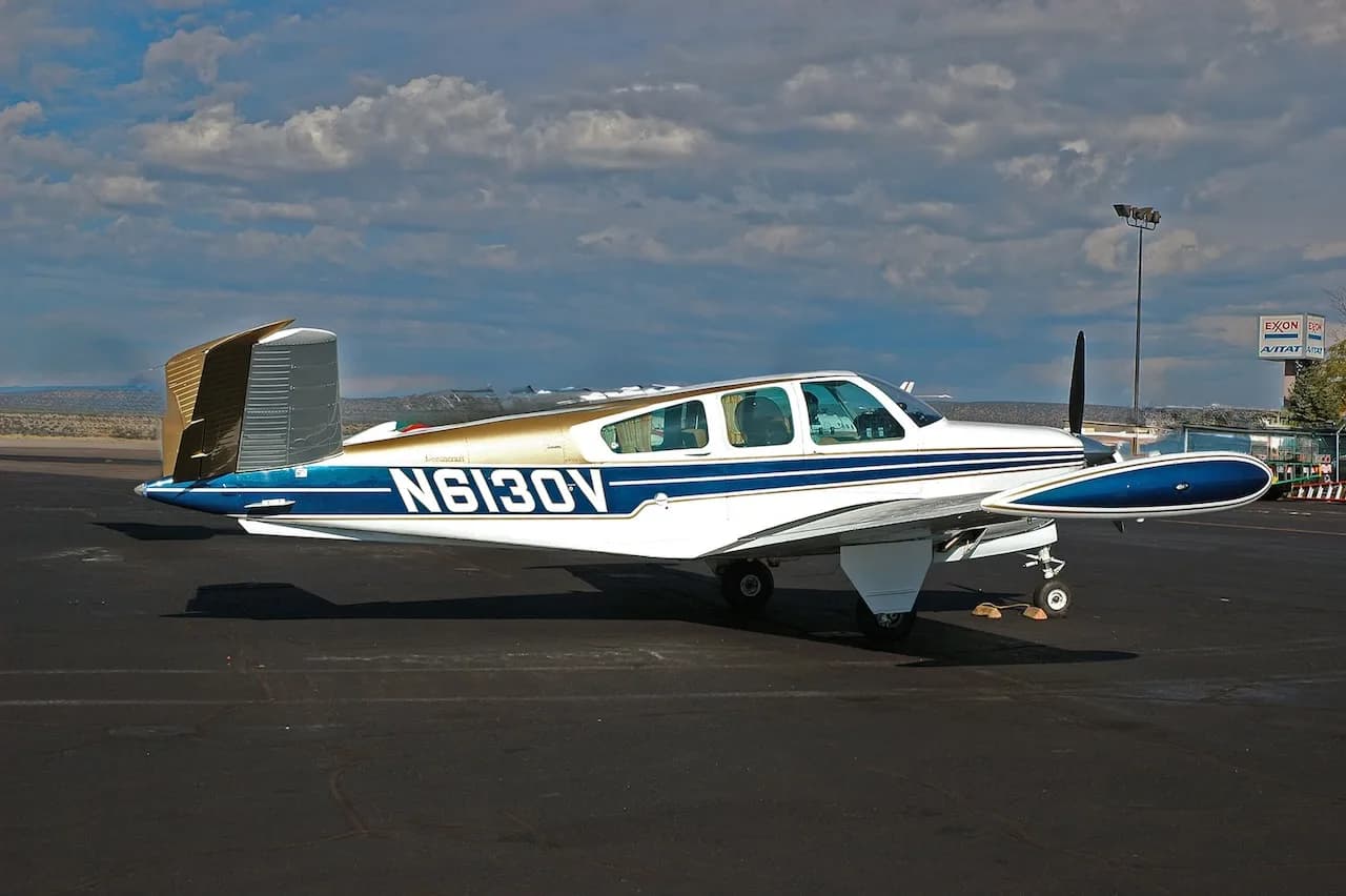 A white, blue, and gold Beechcraft Bonanza N6130V parked on an asphalt ramp under a partly cloudy sky.