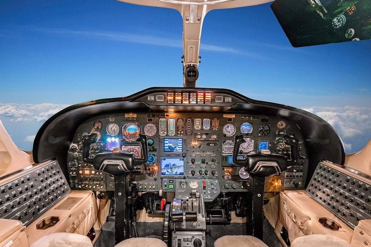 The cockpit of a private jet with multiple screens and controls, showing a view of clouds outside.