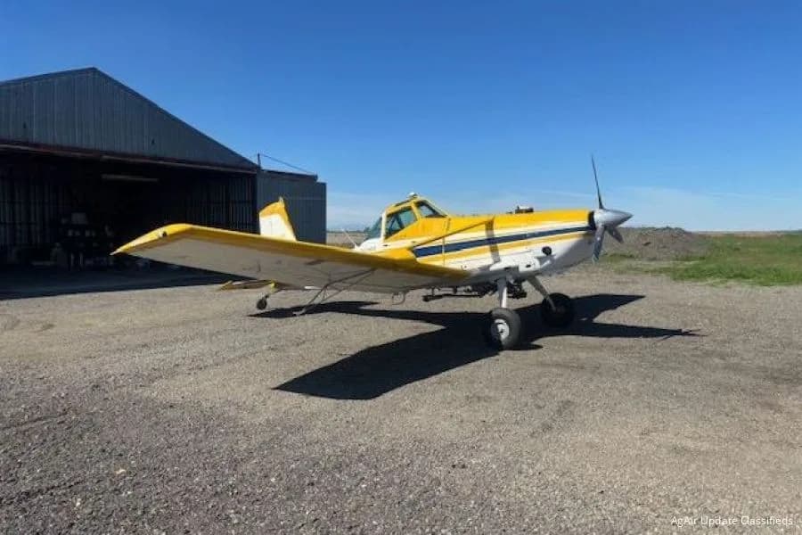 A yellow and white agricultural aircraft with a blue stripe parked on a gravel lot next to a hangar under a clear blue sky.