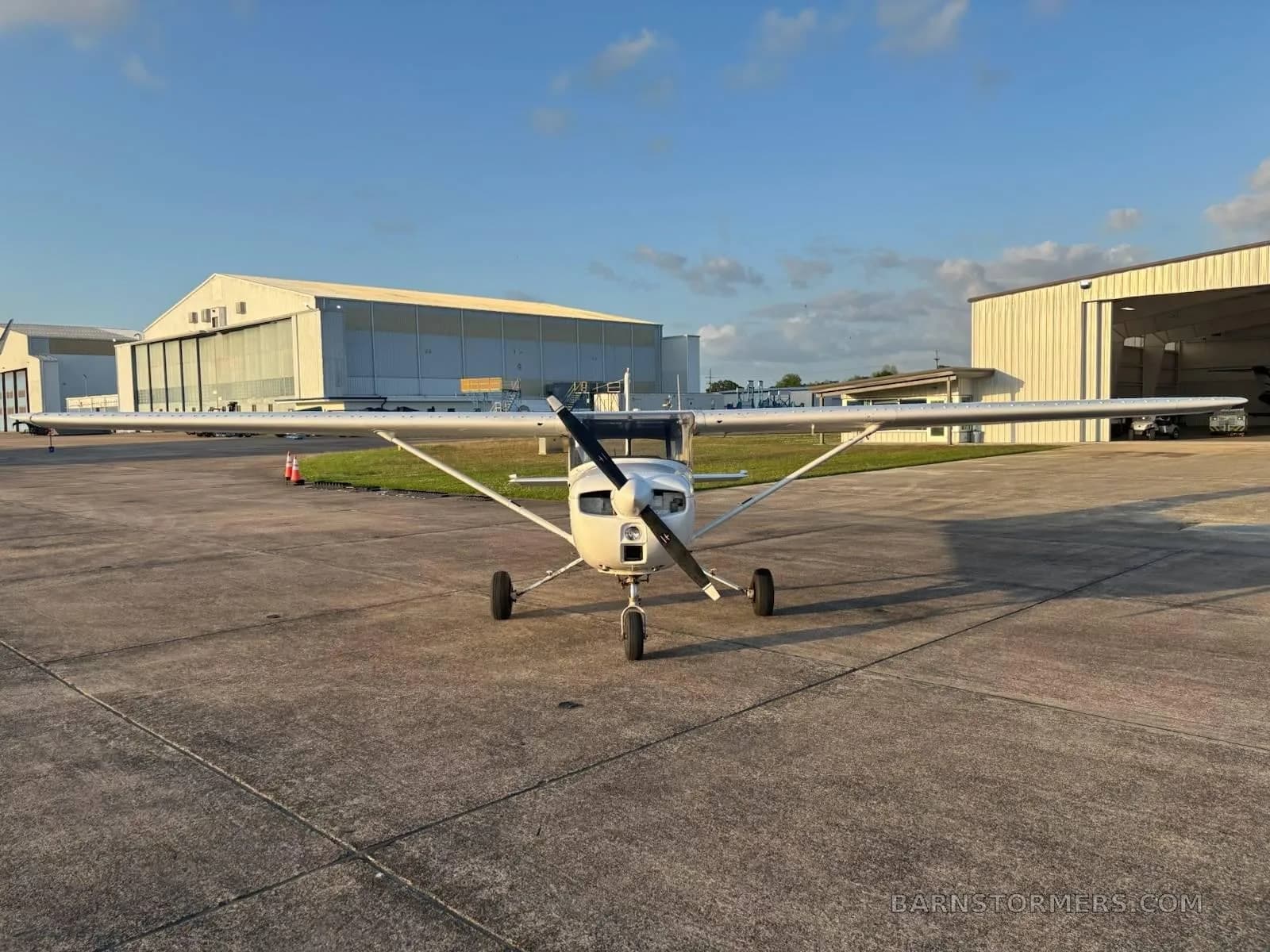 A white Cessna 172 (or similar) is parked head-on on an airfield tarmac with hangars in the background.