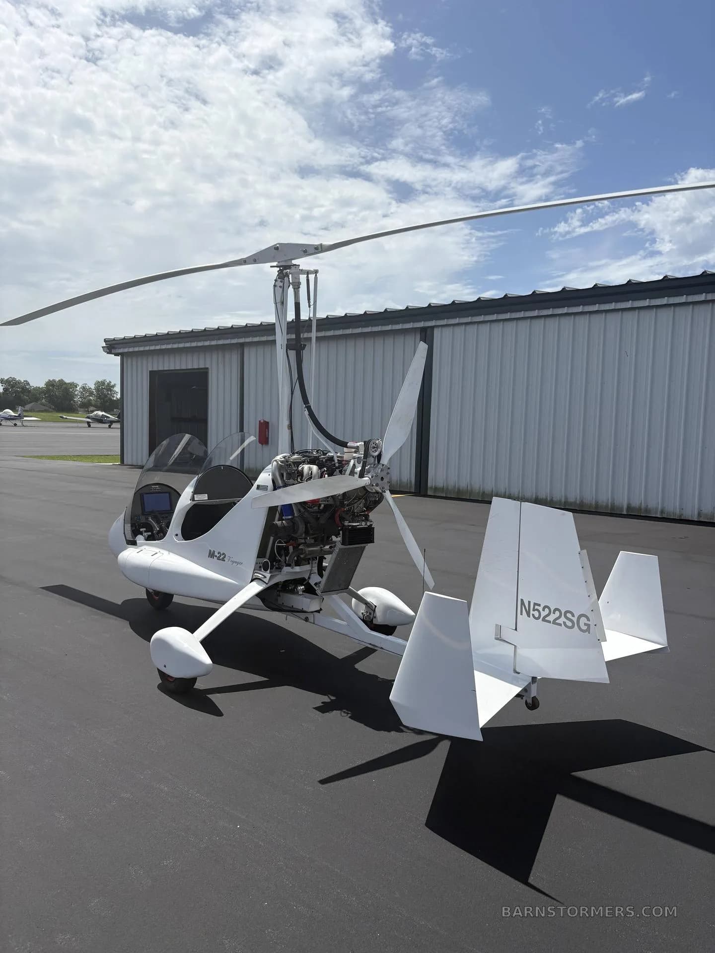 A white M-22 gyrocopter is shown from a rear-quarter view on an asphalt tarmac, highlighting its tail and exposed engine.