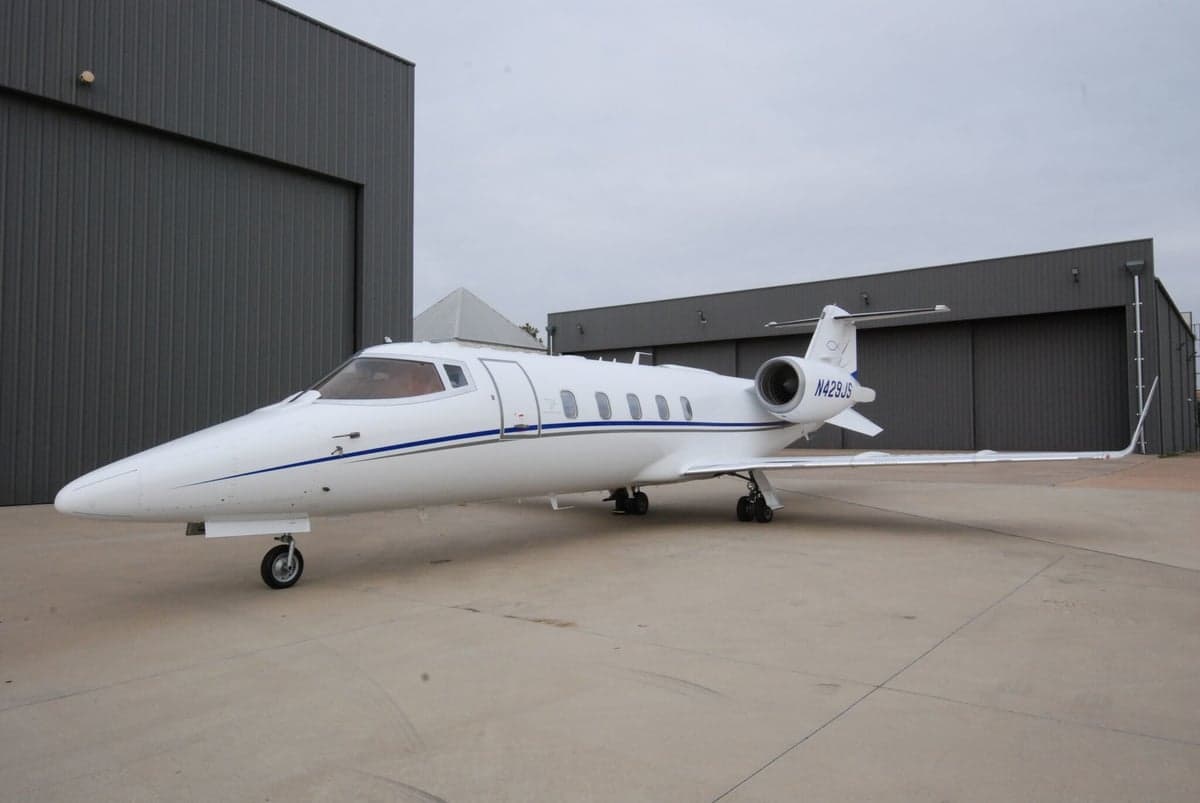 A white business jet with blue stripes and registration N423JS parked on a tarmac in front of dark hangars.