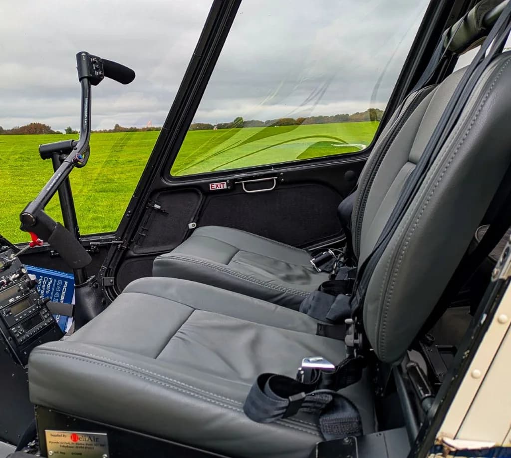 The grey leather interior seats and controls of a helicopter, with a view of a green field through the windows.