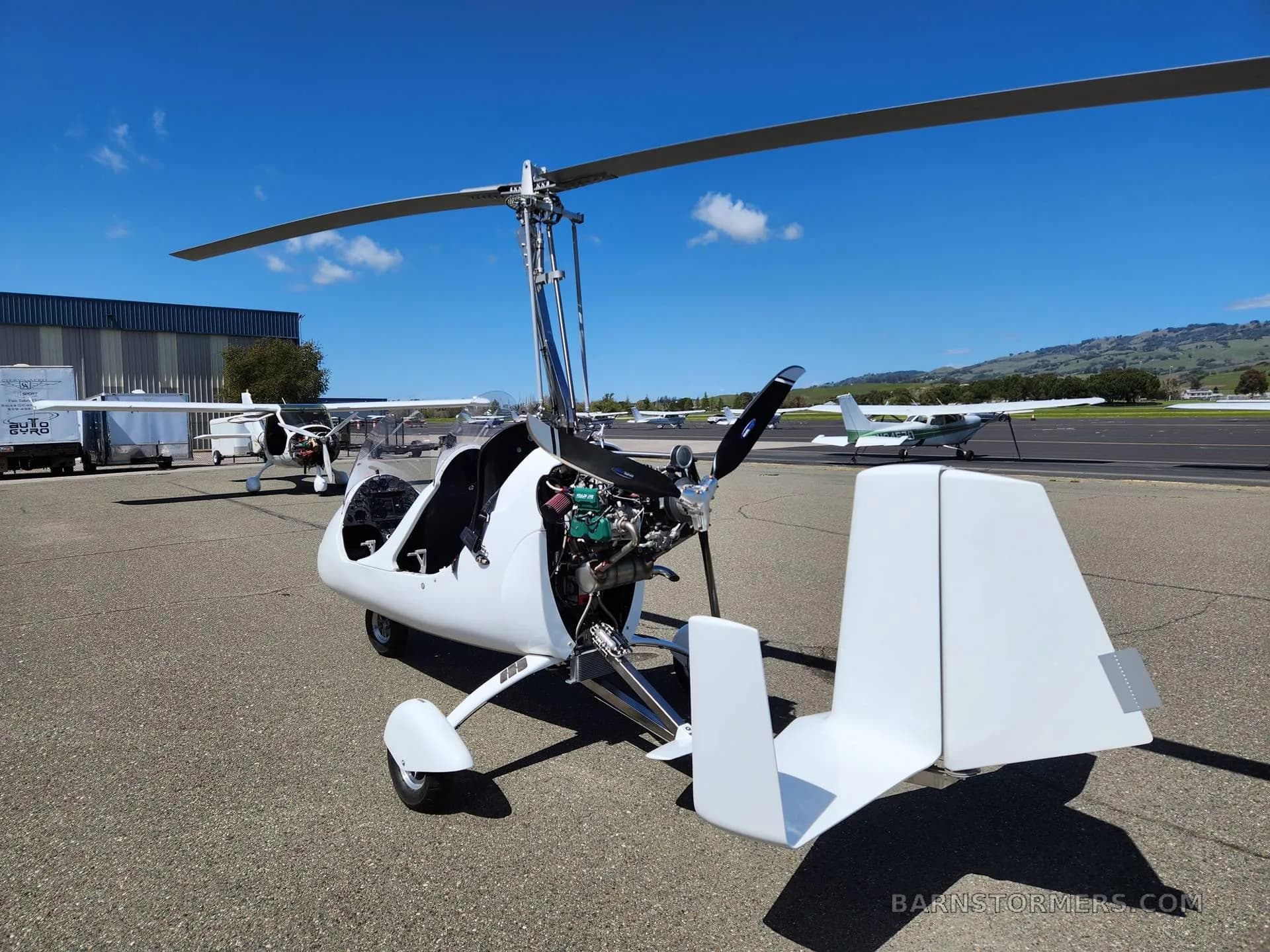 A white gyrocopter with its engine exposed, parked on an airfield tarmac under a clear blue sky.