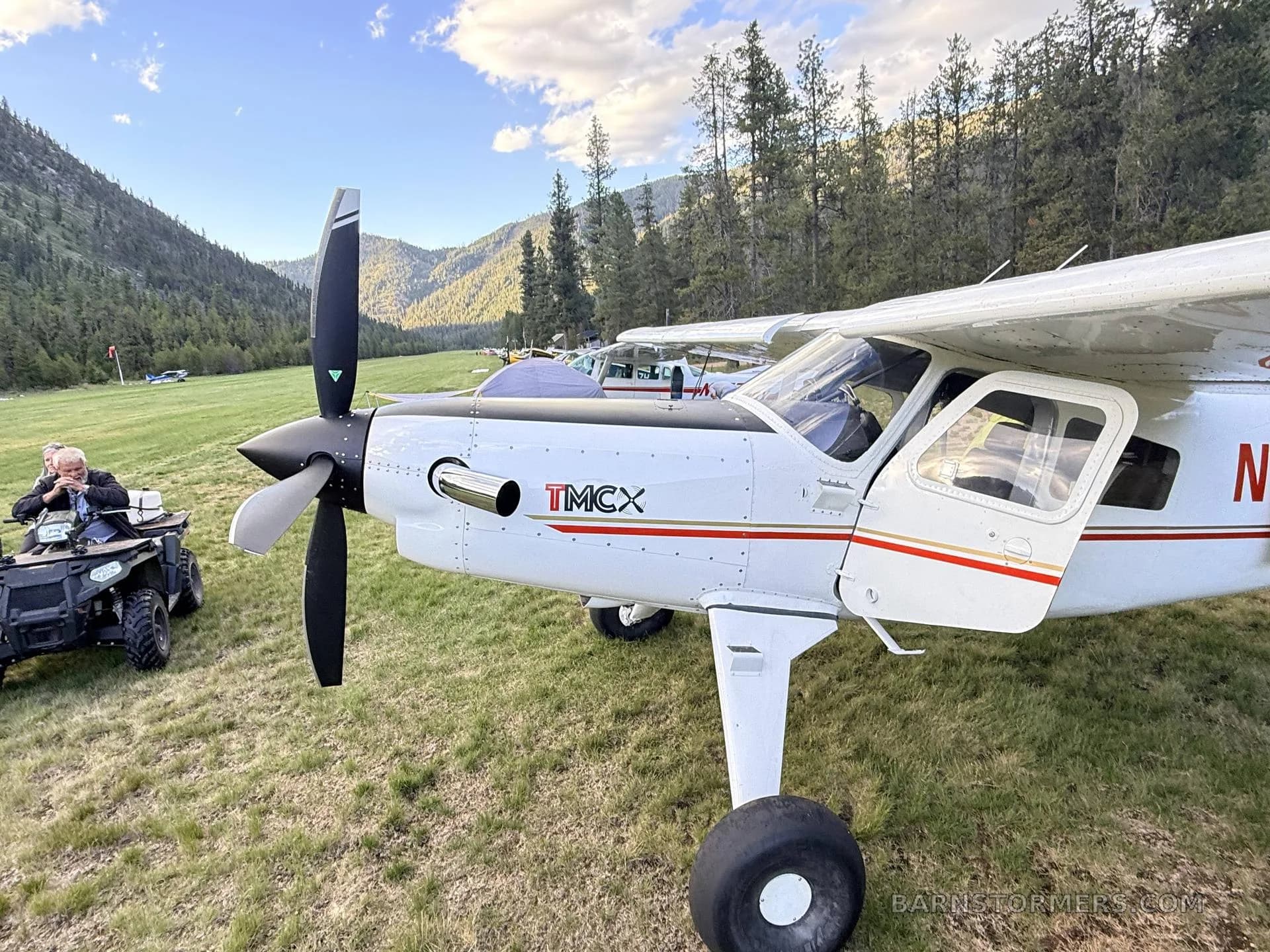 A close-up view of the front of a white and red T-MCX aircraft (N634DV) with its door open, parked on a grassy field with an ATV and people nearby.