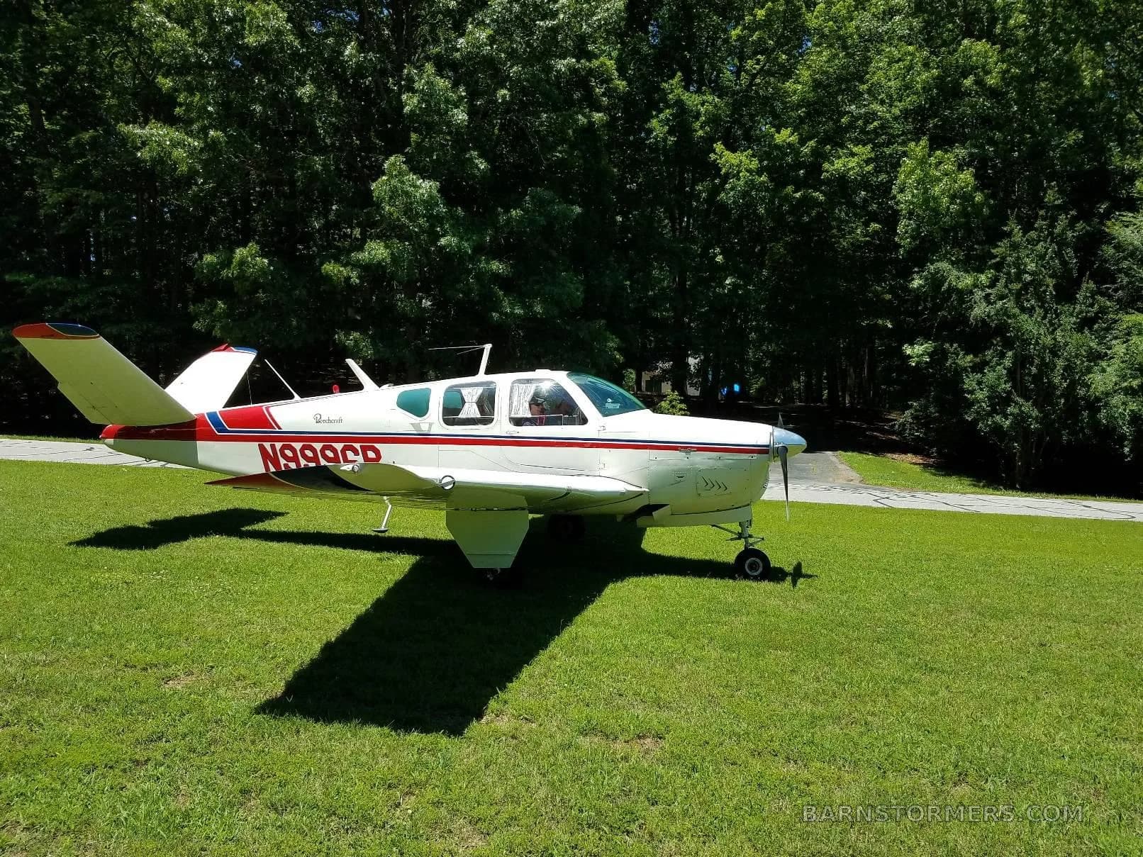 A white Beechcraft Bonanza with red and blue stripes, registration N999CR, parked on a grassy area with trees in the background.