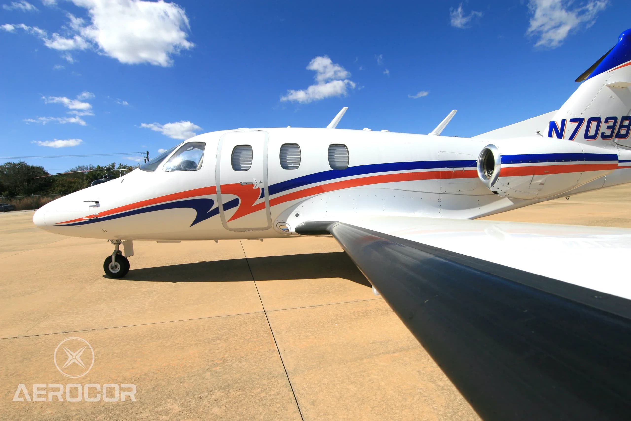 A close-up side view of the front fuselage and wing of a white Eclipse 500 jet with blue and orange stripes, tail number N703BR.