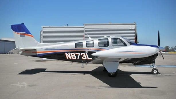 A silver and black single-engine aircraft with blue and orange stripes, tail number N873L, parked on an airfield with a building in the background.