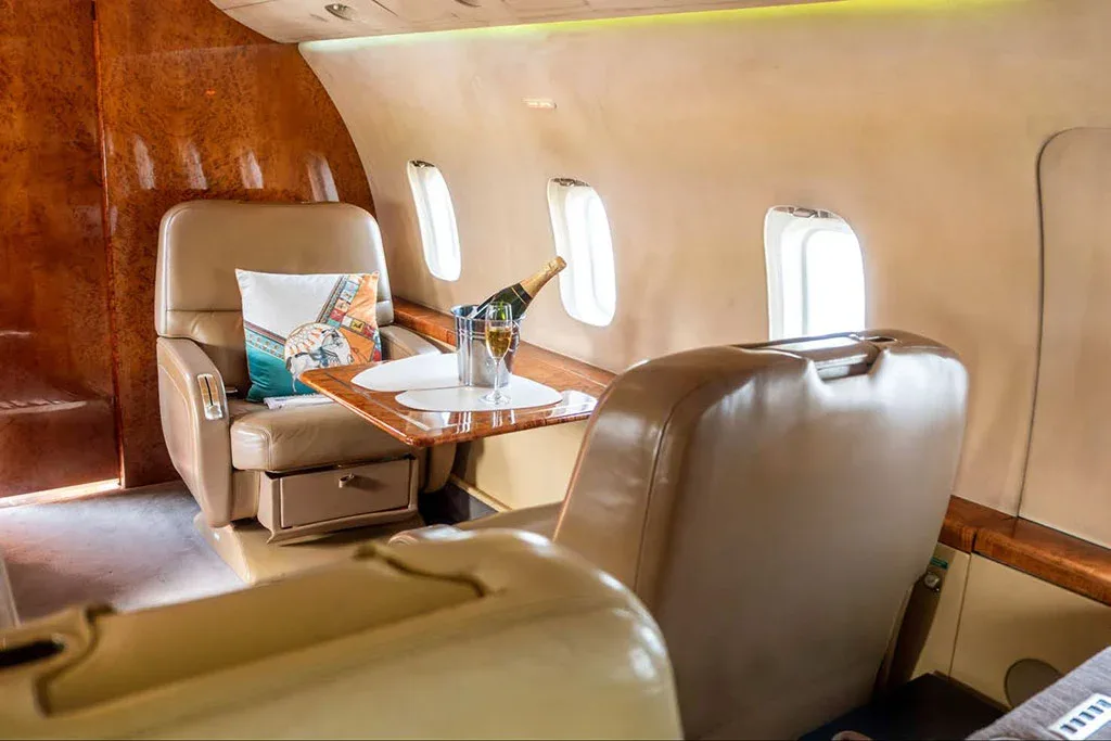 A private jet cabin interior with a tan leather seat, a small table with champagne, and wood paneling.