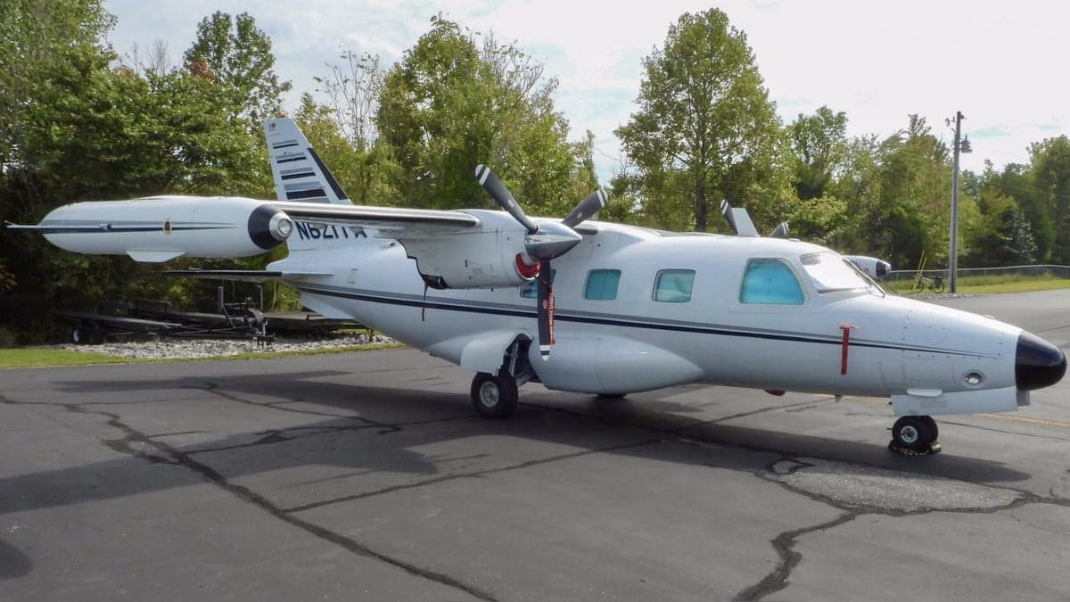 A white Mitsubishi MU-2 aircraft with a black nose, parked on an asphalt ramp with trees in the background.