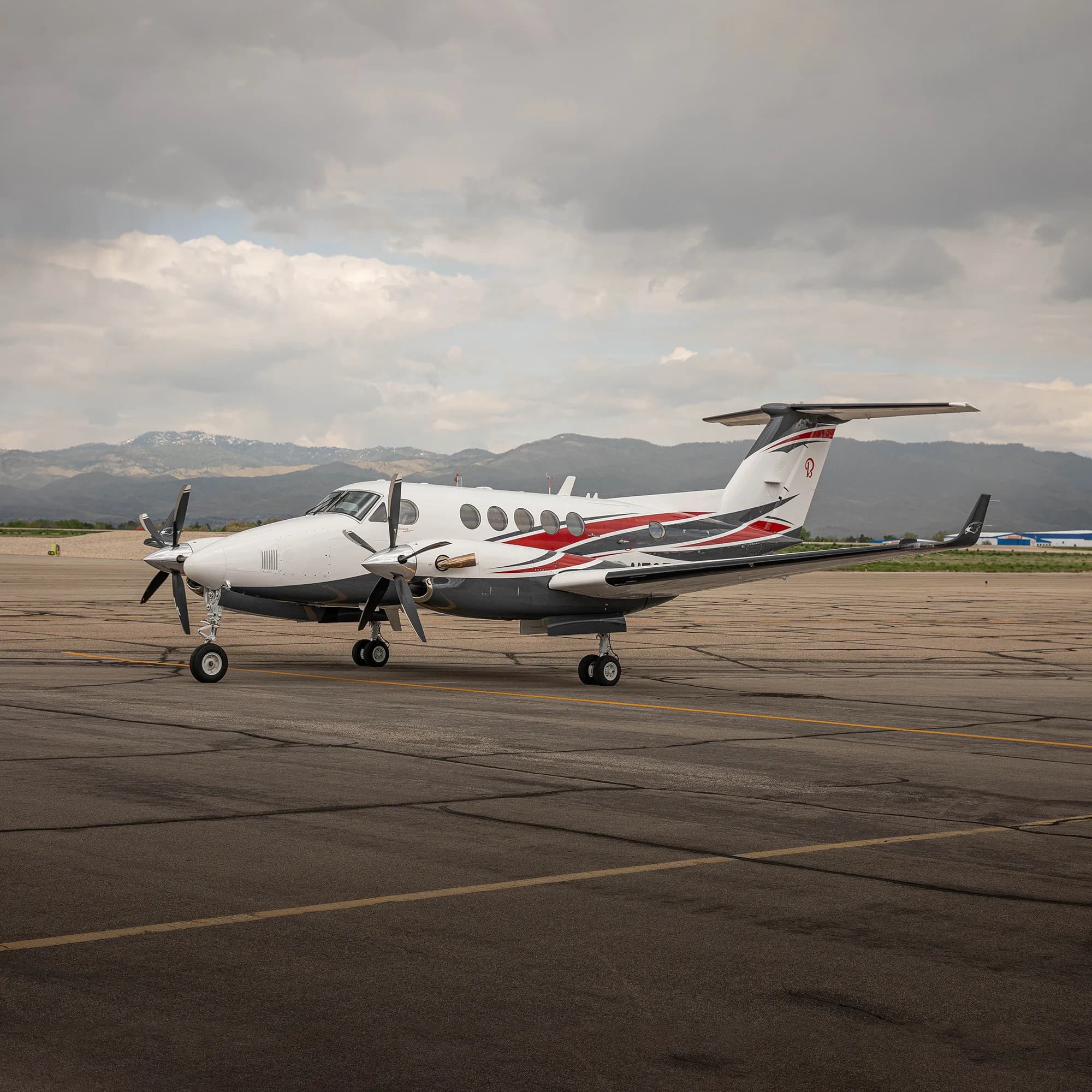 A white, red, and grey twin-turboprop aircraft on an airport tarmac with mountains in the background.