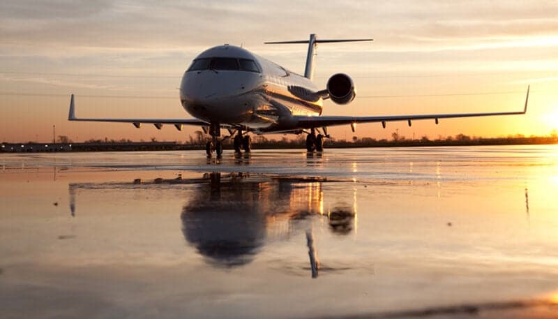 A white private jet with a dark belly is parked on a wet tarmac, reflecting the orange sunset sky.