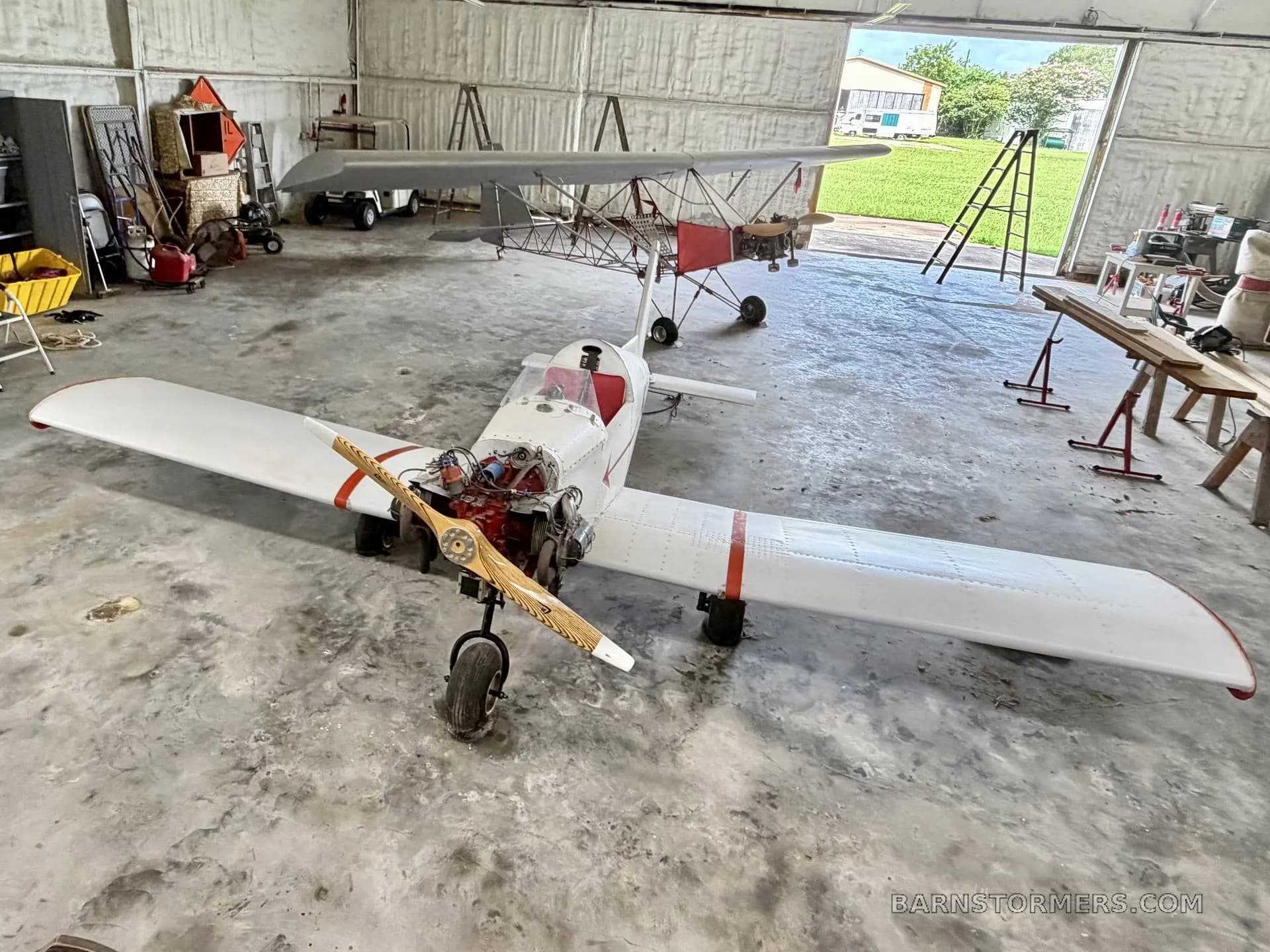 An overhead view of a white single-engine experimental aircraft with a wooden propeller, parked in a hangar.