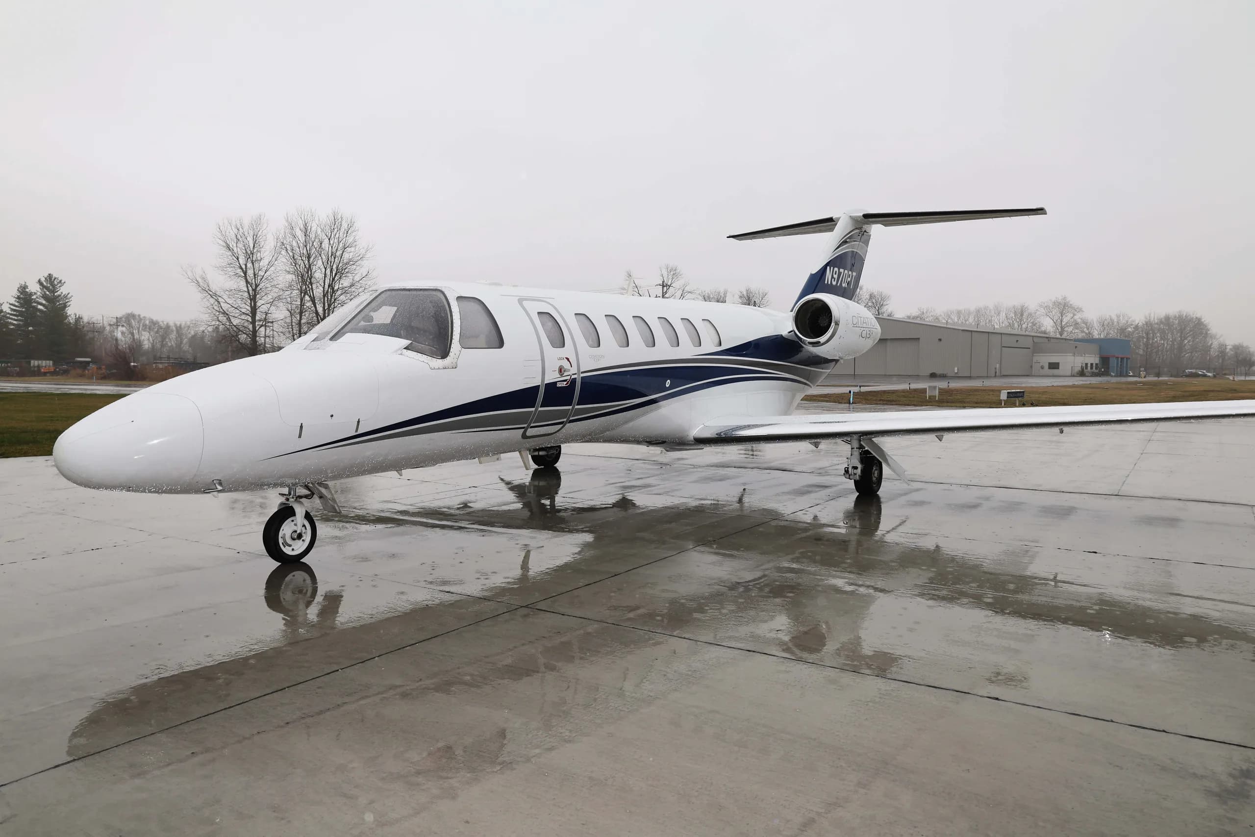 A white Cessna Citation M2 business jet with blue and grey stripes, registration N970PT, parked on a wet tarmac on an overcast day.