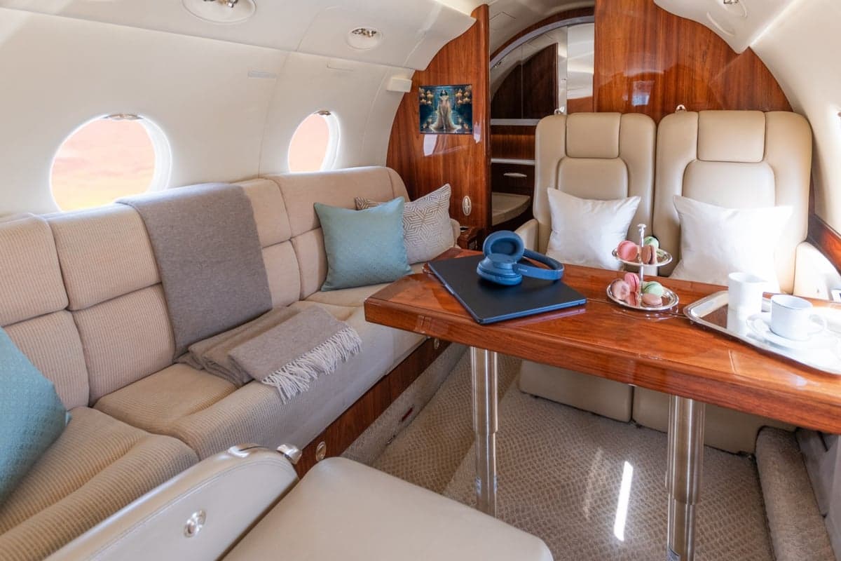 A comfortable aircraft cabin seating area with a beige sofa, a wooden table set with snacks and headphones, and a window view.