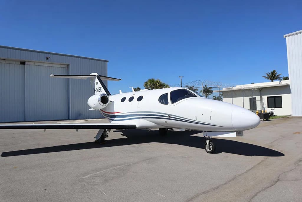A white business jet with blue and red stripes on the tail and fuselage, parked on an asphalt apron in front of a hangar.