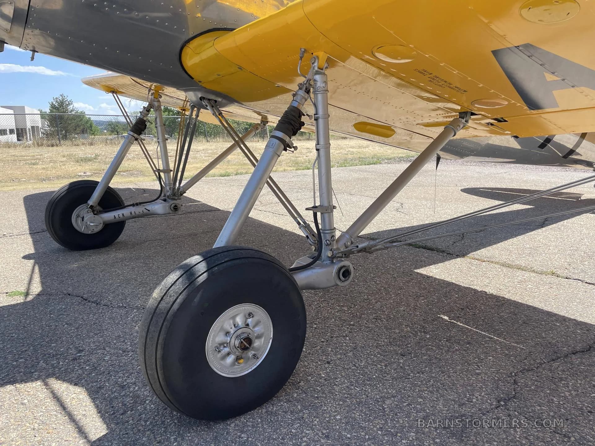 A close-up view of the landing gear and underside of the yellow wing of a biplane on an asphalt surface.