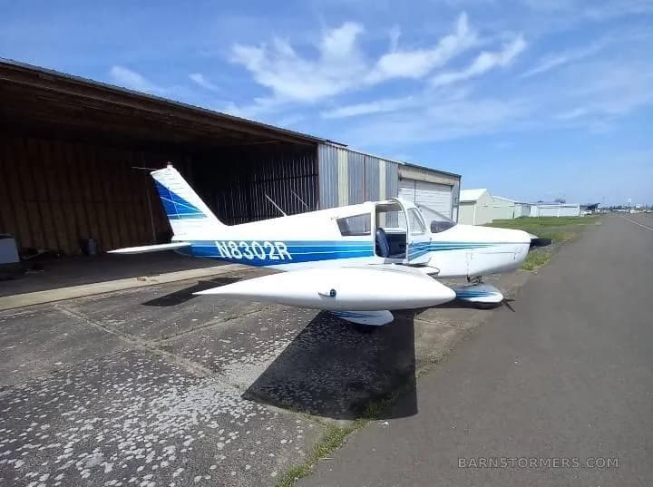 A white and blue Piper Cherokee 140 (N8302R) parked outside a hangar on a sunny day.