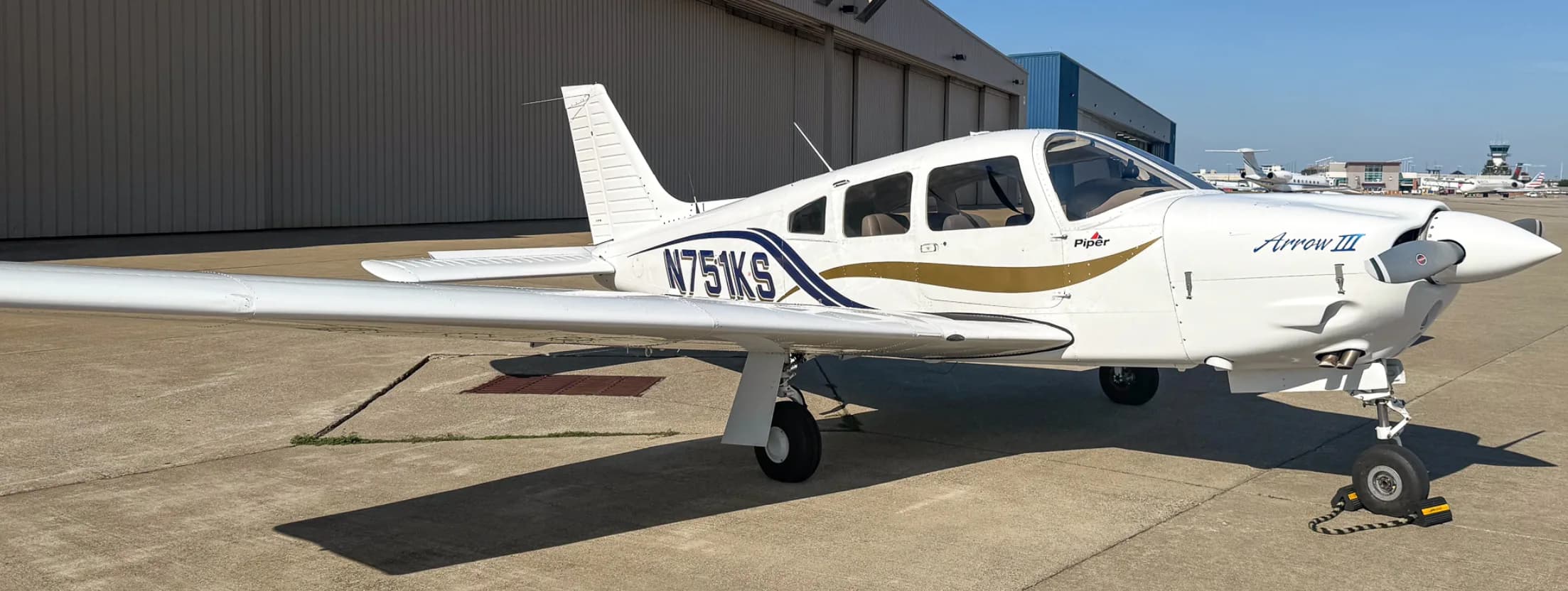 A white Piper Arrow III aircraft with blue and gold stripes, tail number N751KS, parked on an airport tarmac with hangars in the background.