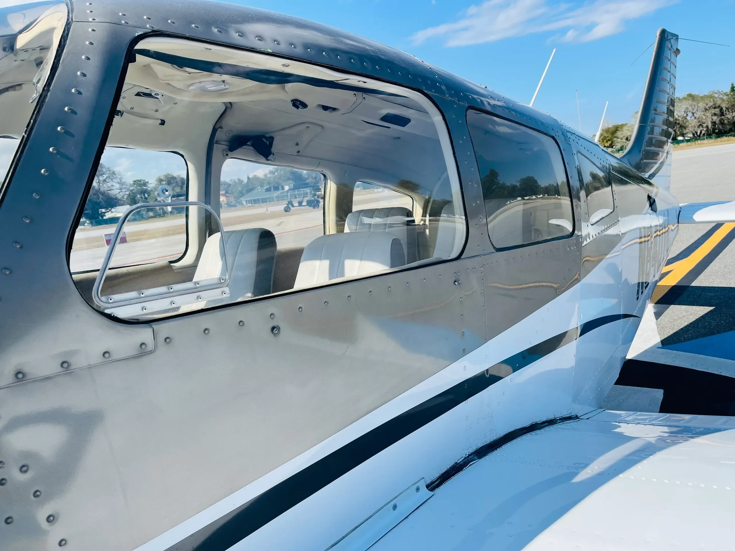 A side exterior view of the cabin windows and fuselage of a gray and white single-engine aircraft on an airfield.