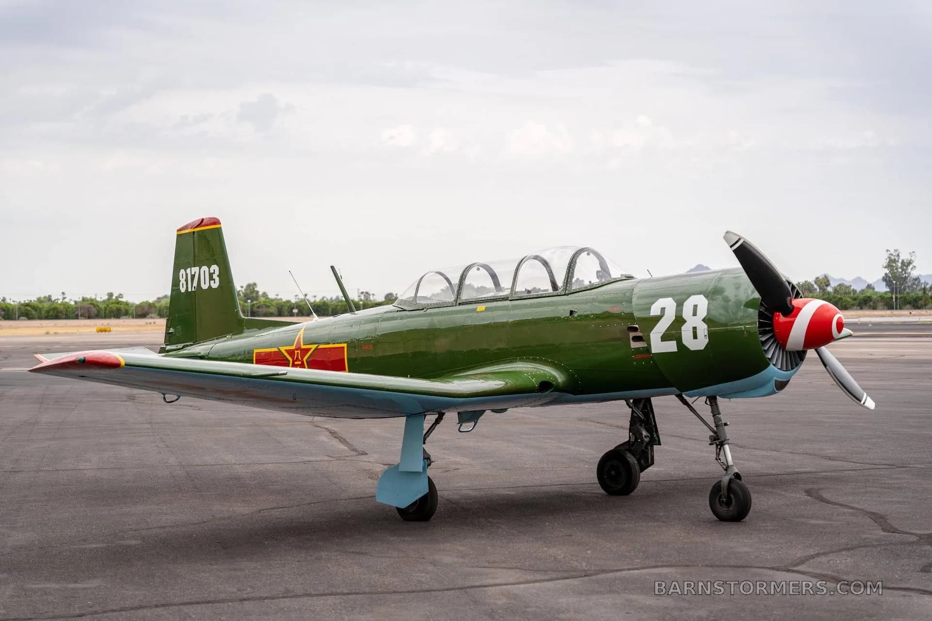 A green Nanchang CJ-6 aircraft with red nose and tail, number 28, parked on an airfield under a cloudy sky.