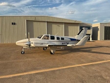 A white and blue Beechcraft Bonanza sits in front of a hangar.