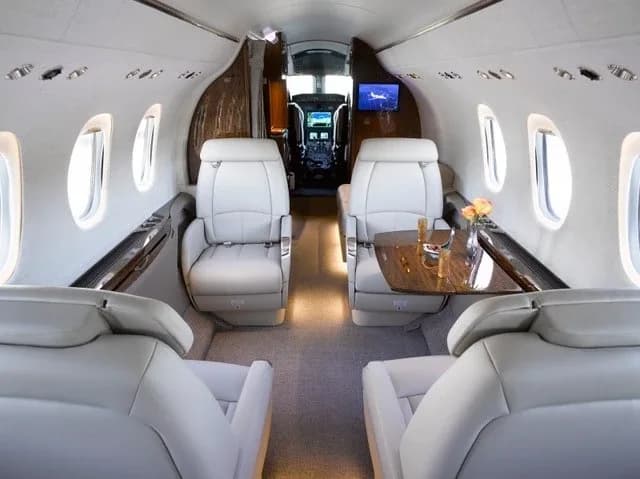 The elegant interior of a business jet cabin, featuring light grey seats, wooden accents, and a view into the cockpit.