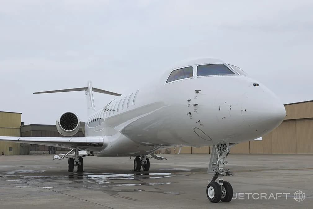 A white Bombardier Global 7500 business jet is parked on a wet tarmac, viewed from the front-right.