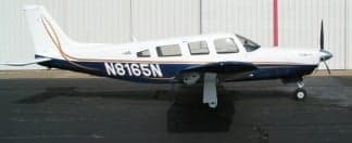 A white and dark blue single-engine propeller aircraft with tail number N8165N is parked on an asphalt tarmac in front of a hangar.