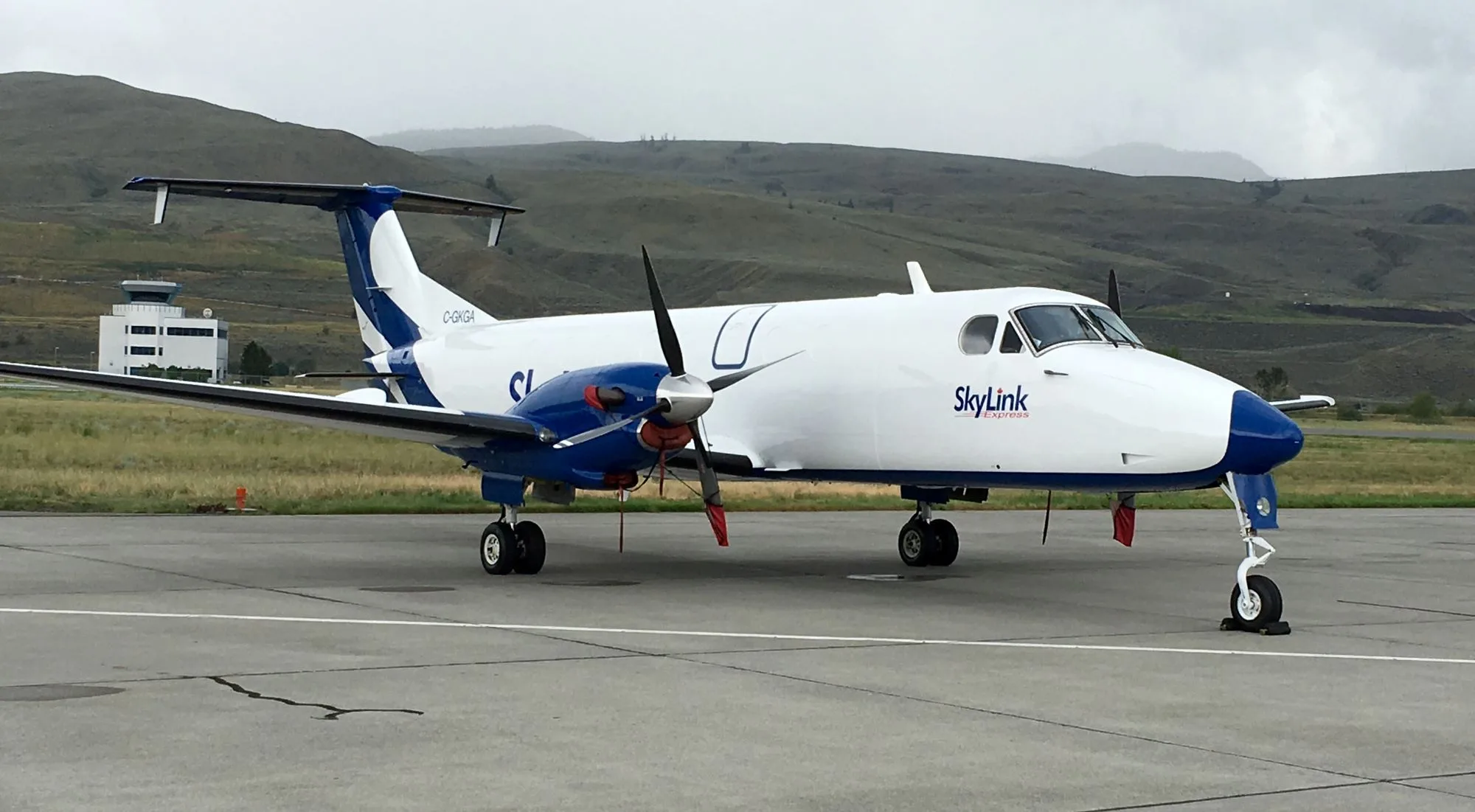 A SkyLink turboprop aircraft parked on an airport tarmac with a control tower and hills in the background.