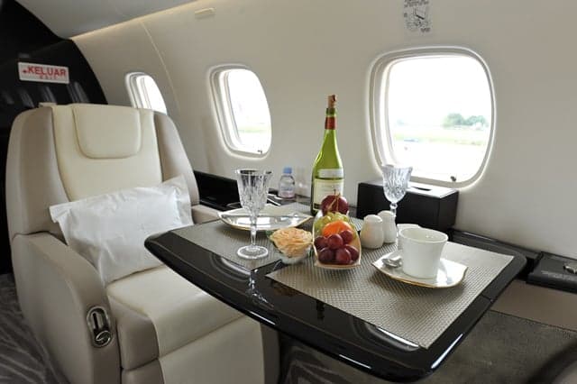 A private jet's interior cabin with a table set with wine, fruit, and glasses next to a cream leather seat.