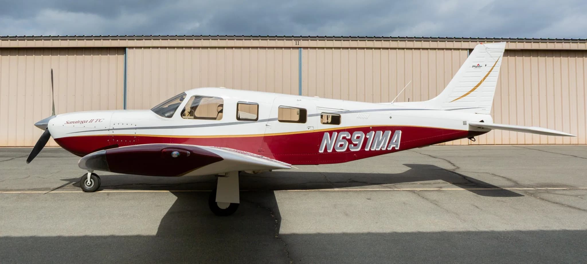 Side view of a white and maroon Piper Saratoga II TC aircraft, registration N691MA, parked in front of a hangar.