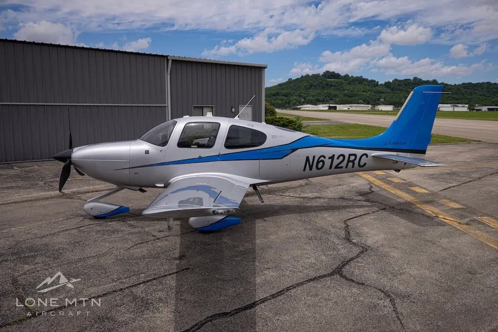 A silver and blue Cirrus SR22T Carbon aircraft, registration N612RC, parked on an airfield next to a hangar.
