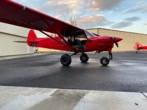 A red Cubcrafters aircraft is parked on a wet runway in front of a hangar.
