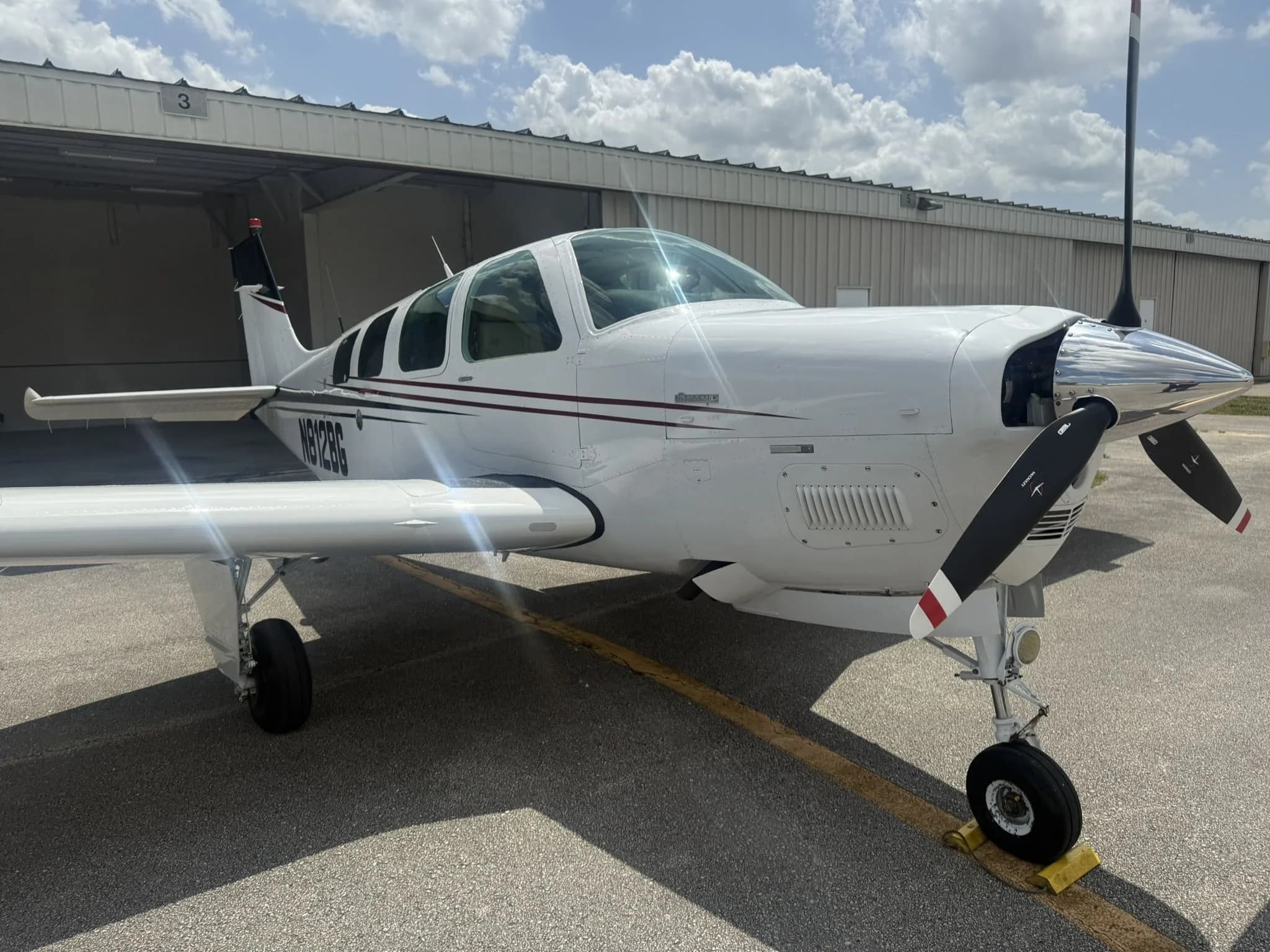 Front view of a white Beechcraft Bonanza N812BG with dark red and black stripes on an airfield.