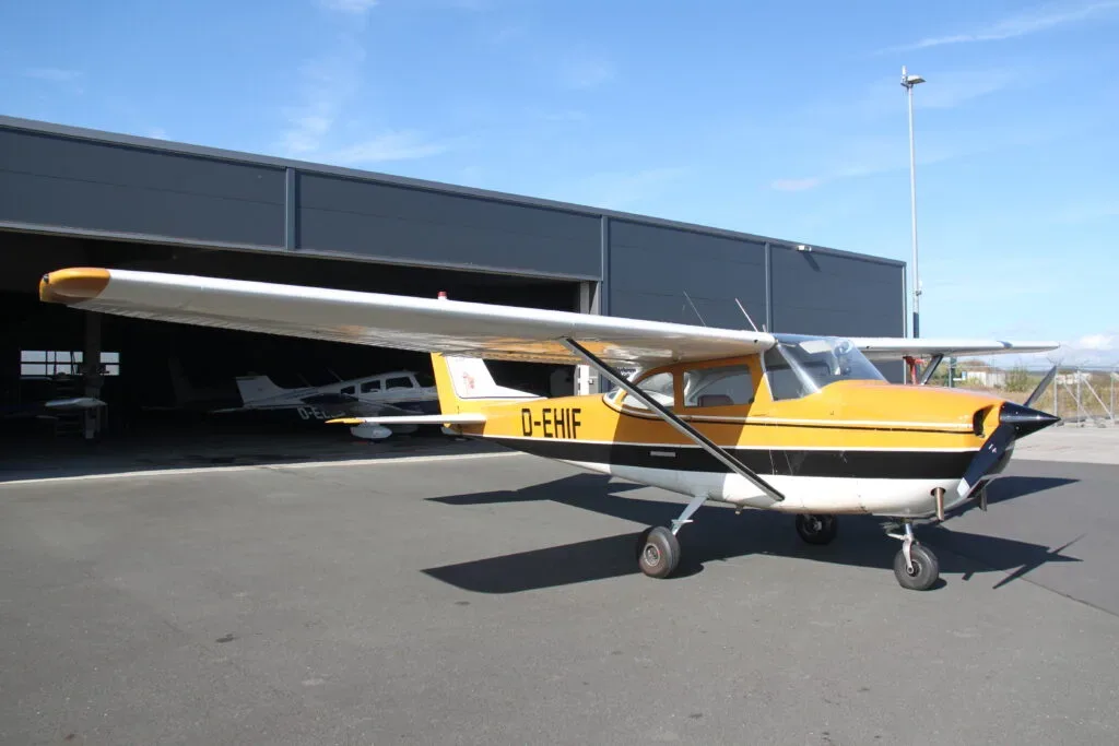 A yellow, black, and white single-engine propeller aircraft is parked on an asphalt apron in front of a hangar on a sunny day.