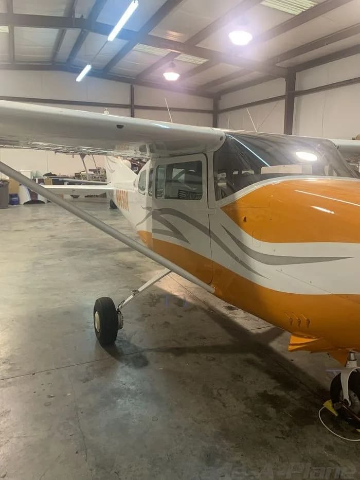 A white high-wing single-engine aircraft with orange and grey stripes, viewed from the side inside a hangar.